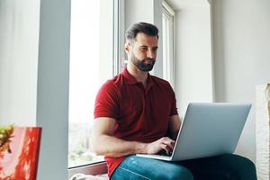 Charming young man in casual clothing using laptop while sitting on the window sill photo