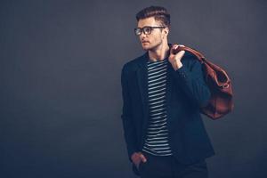 diseñado para el nuevo día. un joven apuesto y seguro de sí mismo con gafas sosteniendo una bolsa y mirando hacia otro lado mientras se enfrenta a un fondo gris foto