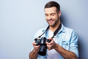 gran tiro guapo joven sosteniendo una cámara digital y mirándola con una sonrisa mientras está de pie contra el fondo gris foto