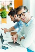 Smart technologies make life easier. Cheerful young man working on digital tablet and looking at camera while man holding mobile phone in the background photo