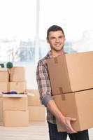 Moving to a new place. Cheerful young man holding a cardboard box and smiling at camera while more carton boxes laying on background photo