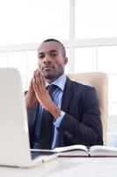 Confident in his power. Confident young African businessman keeping his hands clasped and looking at camera while sitting at his working place photo