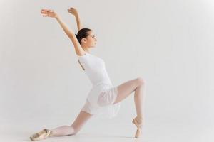 gracia en cada movimiento. Vista lateral de la hermosa bailarina joven en tutú blanco bailando contra el fondo blanco. foto