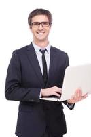 Quality support for your business. Handsome young man in formalwear working on laptop and smiling while standing isolated on white background photo