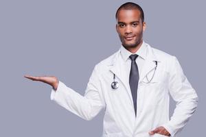 Advertising your product. Confident African doctor holding copy space and looking at camera while standing against grey background photo