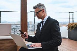 Confident mature man working on laptop and using mobile phone while standing on the rooftop terrace photo