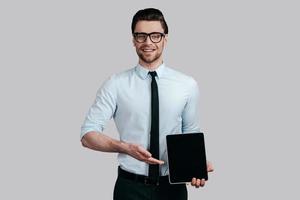 Copy space on his tablet.  Handsome young man in white shirt and tie pointing copy space on his digital tablet and looking at camera while standing against grey background photo