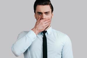 Trying to stop gossiping. Surprised young man in white shirt and tie covering mouth with hand and looking at camera while standing against grey background photo