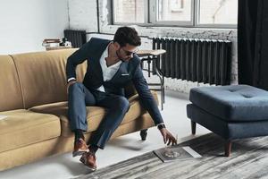 Time to read. Good looking young man in full suit taking magazine from the floor while sitting on the sofa photo