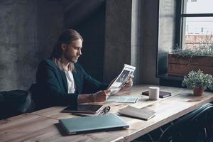 leyendo las últimas noticias. un joven apuesto con ropa informal inteligente leyendo un periódico mientras se sienta en su escritorio en la oficina foto