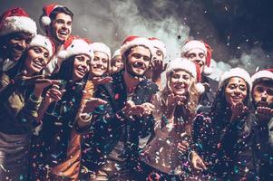 poca magia navideña. grupo de hermosos jóvenes con sombreros de santa que soplan confeti colorido y se ven felices foto