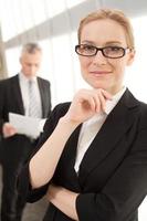 Confident and intelligent. Attractive mature businesswoman holding hand on chin and smiling at camera while man in formalwear standing on background photo