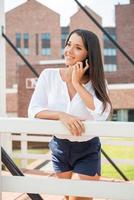 Talking with friend. Beautiful young woman talking on the mobile phone and smiling while standing outdoors photo