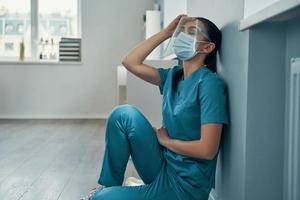 Tired young female nurse in protective workwear touching her head while working in the hospital photo