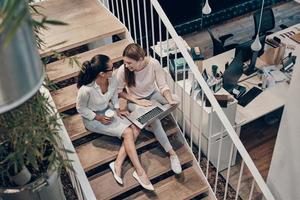 Top view of two young modern women in smart casual wear communicating and using laptop while sitting on the stairs photo