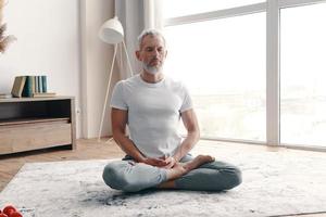 hombre mayor con ropa deportiva meditando mientras se sienta en el suelo en la posición de loto en casa foto