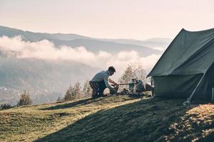 Cooking breakfast. Young man making a campfire while sitting near the tent in the mountains photo