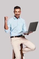 Finally success Good looking young man smiling and gesturing while sitting on stool against grey background photo