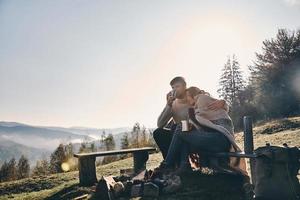 Appreciating every minute together. Beautiful young couple having morning coffee while sitting by the campfire in mountains photo