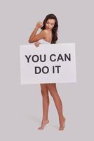 Time to be brave.  Full length of attractive young woman covering with a poster and looking at camera with a smile while standing against grey background photo