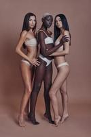 feminidad. Longitud total de tres atractivas mujeres de raza mixta mirando a la cámara mientras están de pie contra el fondo marrón foto