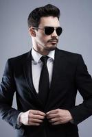 Making business look good. Handsome young man in formalwear and sunglasses buttoning his jacket and looking away while standing against grey background photo