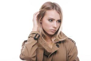 Trendy beauty. Attractive young woman in coat holding hand in hair and looking away while standing against white background photo