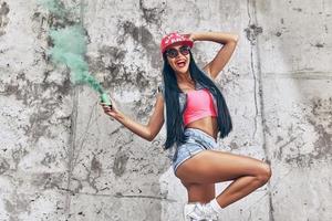 diversión de colores vista de ángulo bajo de mujeres jóvenes alegres sosteniendo una bomba de humo y sonriendo mientras posan contra la pared de hormigón foto