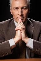 Thinking about new solutions. Thoughtful senior man in formalwear sitting at the table and holding hands clasped while isolated on black background photo