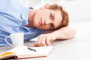 descansando en casa. un joven apuesto tirado en el suelo y mirando la cámara con un bloc de notas y una taza de café cerca de él foto