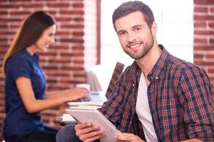 Confident and creative. Handsome young man holding digital tablet and smiling while woman sitting in the background and working on laptop photo