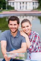 viajeros felices de la ciudad. vista superior de la feliz pareja de jóvenes turistas sentados cerca de un hermoso edificio y mirando a la cámara mientras el hombre sostiene el mapa foto