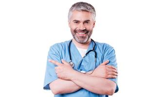 Doctor pointing copy space. Cheerful mature surgeon pointing away and smiling while keeping arms crossed and standing isolated on white photo