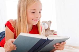 leyendo su libro favorito. linda niña leyendo un libro y sonriendo mientras está acostada en la cama foto