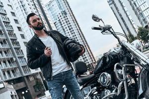 Do you want to take a ride Handsome young man in leather jacket looking away while standing near the motorbike outdoors photo
