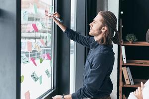 trabajar algunos conceptos. joven moderno con ropa informal inteligente usando notas adhesivas en la ventana mientras trabaja con su colega en la oficina foto