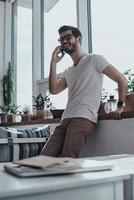 Always in touch with friends. Handsome young man talking on his smart phone while leaning on the window sill photo