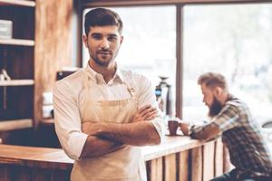 Loving his work. Young handsome man in apron looking at camera and keeping arms crossed while standing at cafe with customer at background photo