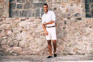 confiado en su estilo perfecto. toda la longitud de un joven africano alegre con ropa informal elegante que se ajusta la manga de la camisa y mira la cámara mientras está de pie contra la pared de piedra al aire libre foto