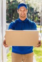 Take your package Happy young delivery man stretching out a cardboard box while standing at the entrance of apartment photo