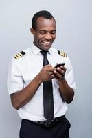 Messages after landing. Side view of happy African pilot in uniform holding mobile phone while standing against grey background photo