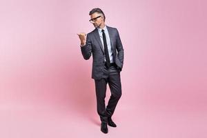 Confident mature man in full suit pointing away while standing against pink background photo