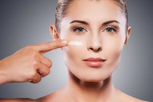 manteniendo su piel joven y suave. retrato de una mujer madura sin camisa esparciendo crema en la cara y sonriendo mientras se enfrenta a un fondo gris foto