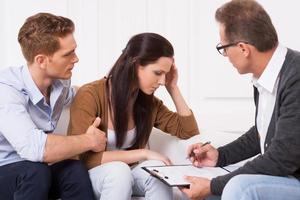 Feeling hopeless. Young man consoling his depressed wife while sitting together with psychiatrist photo