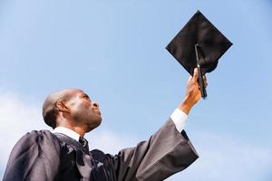 feliz graduado. vista en ángulo bajo de un joven africano feliz con un vestido de graduación sosteniendo su tablero de mortero contra el cielo azul foto