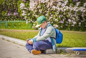 A young man is sitting on the side of a lilac alley and looking at his phone.  Against the background of lilac bushes. Interactions. Selective focus. photo