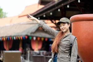 joven viajera asiática con máscara y mochila señalando con el dedo las famosas atracciones turísticas de tailandia foto