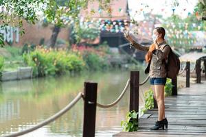 Young asian woman traveler with mask and backpack using mobile phone taking selfie while solo travel in summer sunny day. photo