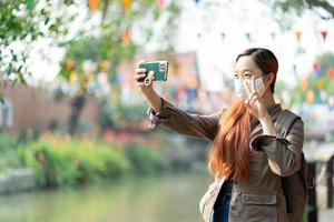 Hipster woman traveler with mask and backpack using mobile phone taking selfie while solo travel in summer sunny day. photo