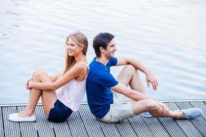 They love spending time together. Beautiful young loving couple sitting back to back at the quayside and looking away with smile photo